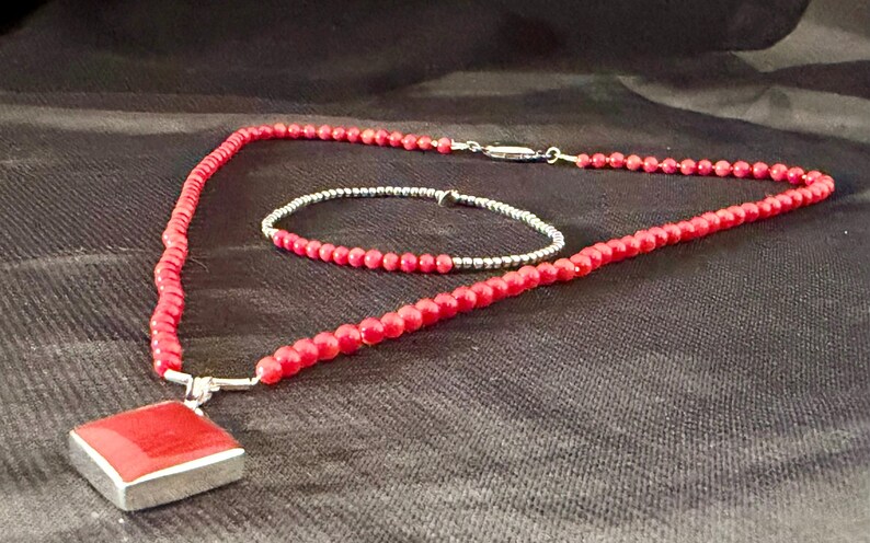 May include: A red beaded necklace with a square pendant and a matching bracelet. The pendant is a square of red material set in a silver frame. The necklace and bracelet are made of small, round red beads. The jewelry is displayed on a dark background.