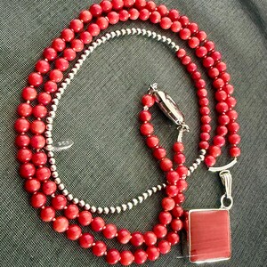 May include: A layered necklace featuring red coral beads and a strand of small silver beads. The necklace has a silver clasp and a square red pendant. The jewelry is displayed on a gray surface.