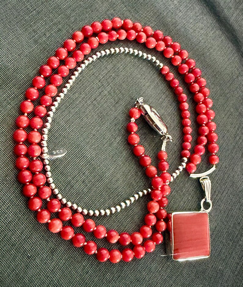 May include: A layered necklace featuring red coral beads and a strand of small silver beads. The necklace has a silver clasp and a square red pendant. The jewelry is displayed on a gray surface.
