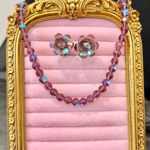 May include: A vintage jewelry set featuring a necklace and matching earrings. The set includes a beaded necklace and flower-shaped earrings, all in a deep purple hue with iridescent accents. The jewelry is displayed on a pink velvet surface within a gold-framed display.
