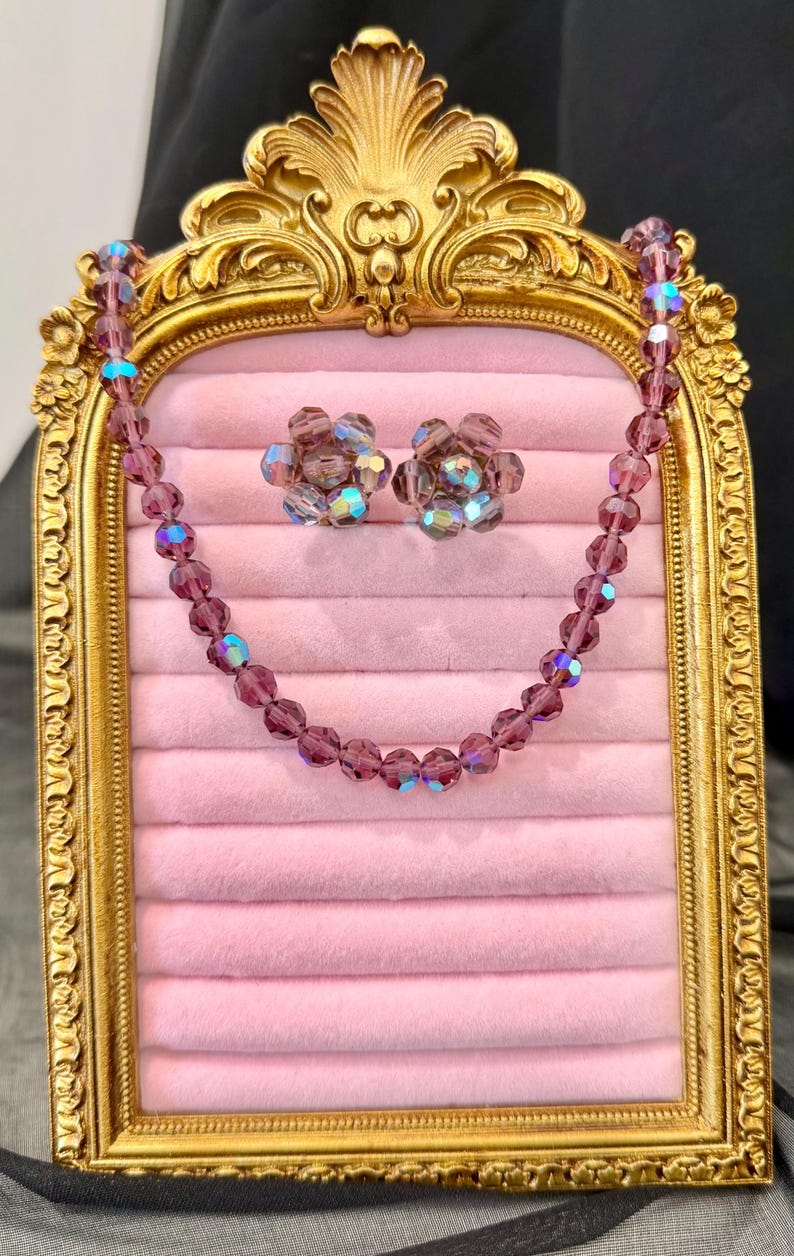 May include: A vintage jewelry set featuring a necklace and matching earrings. The set includes a beaded necklace and flower-shaped earrings, all in a deep purple hue with iridescent accents. The jewelry is displayed on a pink velvet surface within a gold-framed display.
