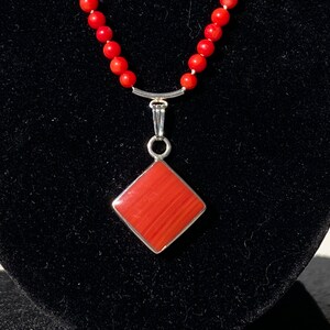 May include: A red coral bead necklace with a silver pendant. The pendant is a square shape with a red, textured surface and a silver border. The necklace is displayed on a black velvet surface.