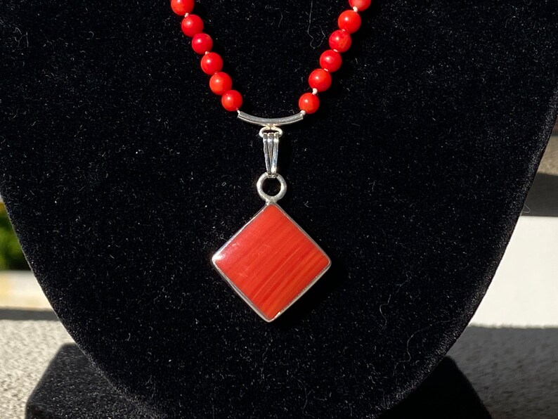 May include: A red coral bead necklace with a silver pendant. The pendant is a square shape with a red, textured surface and a silver border. The necklace is displayed on a black velvet surface.