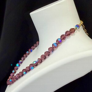 May include: A purple beaded necklace displayed on a white mannequin. The necklace features faceted beads with iridescent highlights. The clasp and chain are gold-colored. The background is black.