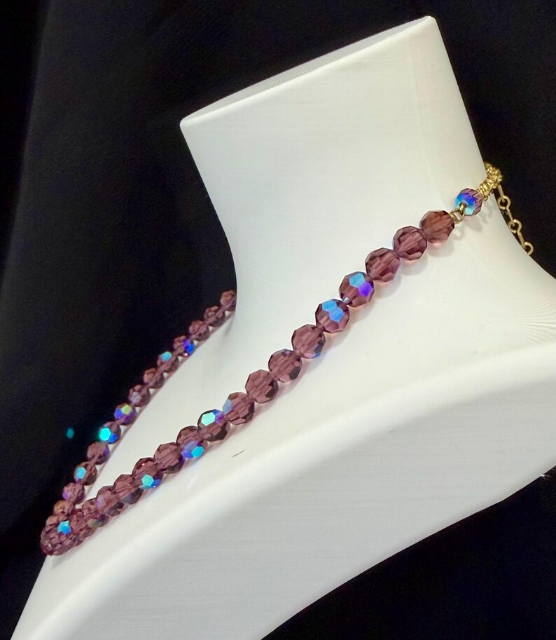 May include: A purple beaded necklace displayed on a white mannequin. The necklace features faceted beads with iridescent highlights. The clasp and chain are gold-colored. The background is black.