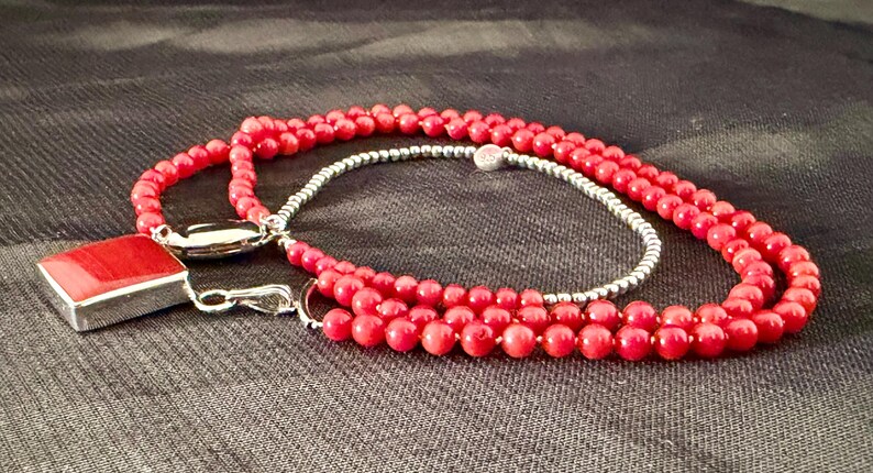 May include: A red beaded necklace with a silver clasp and a square pendant. The necklace has a double strand of small, round beads and a single strand of silver beads. The pendant is a red square set in a silver frame.