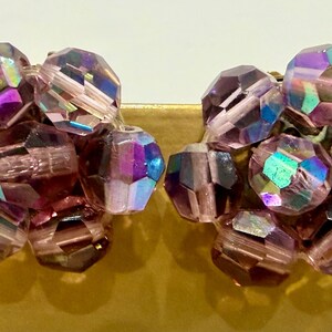 May include: A pair of vintage clip-on earrings featuring clusters of faceted, iridescent beads. The beads are primarily shades of purple and pink, with flashes of blue and green. The earrings have a gold-tone metal clip.