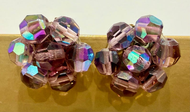 May include: A pair of vintage clip-on earrings featuring clusters of faceted, iridescent beads. The beads are primarily shades of purple and pink, with flashes of blue and green. The earrings have a gold-tone metal clip.