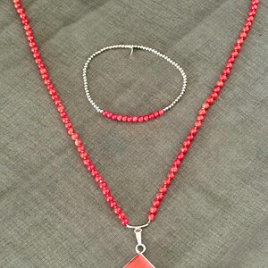May include: A red beaded necklace with a silver pendant and a matching bracelet. The necklace features a diamond-shaped pendant. The jewelry is displayed on a gray surface.