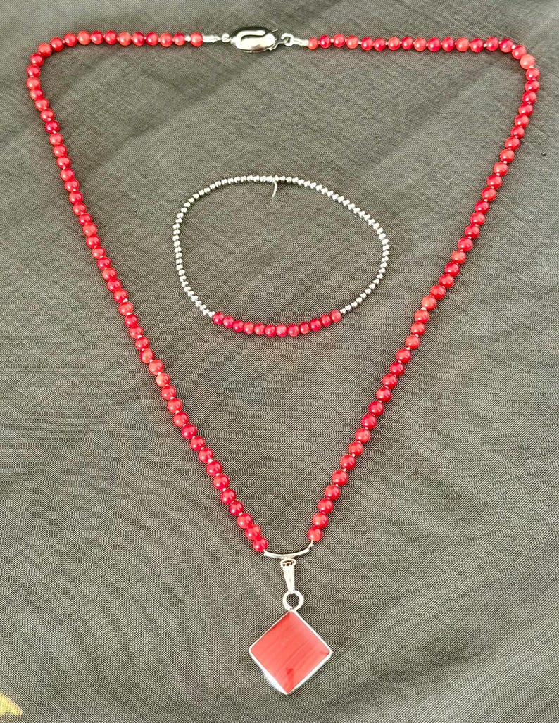 May include: A red beaded necklace with a silver pendant and a matching bracelet. The necklace features a diamond-shaped pendant. The jewelry is displayed on a gray surface.