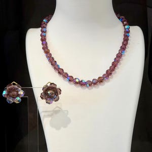 May include: A vintage jewelry set featuring a beaded necklace and matching clip-on earrings. The necklace and earrings are a deep purple color with iridescent accents. The earrings are flower-shaped. The jewelry is displayed on a white mannequin.