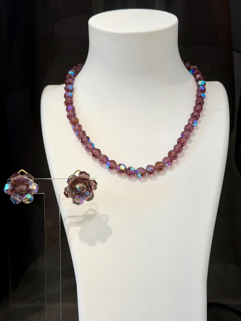 May include: A vintage jewelry set featuring a beaded necklace and matching clip-on earrings. The necklace and earrings are a deep purple color with iridescent accents. The earrings are flower-shaped. The jewelry is displayed on a white mannequin.