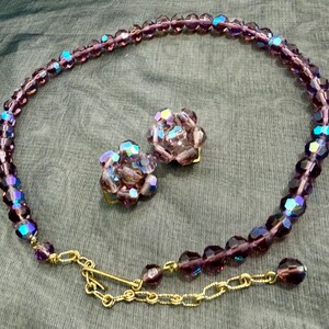 May include: A vintage jewelry set featuring a necklace and matching clip-on earrings. The necklace and earrings are made of faceted, iridescent purple beads. The necklace has a gold-tone chain and clasp.