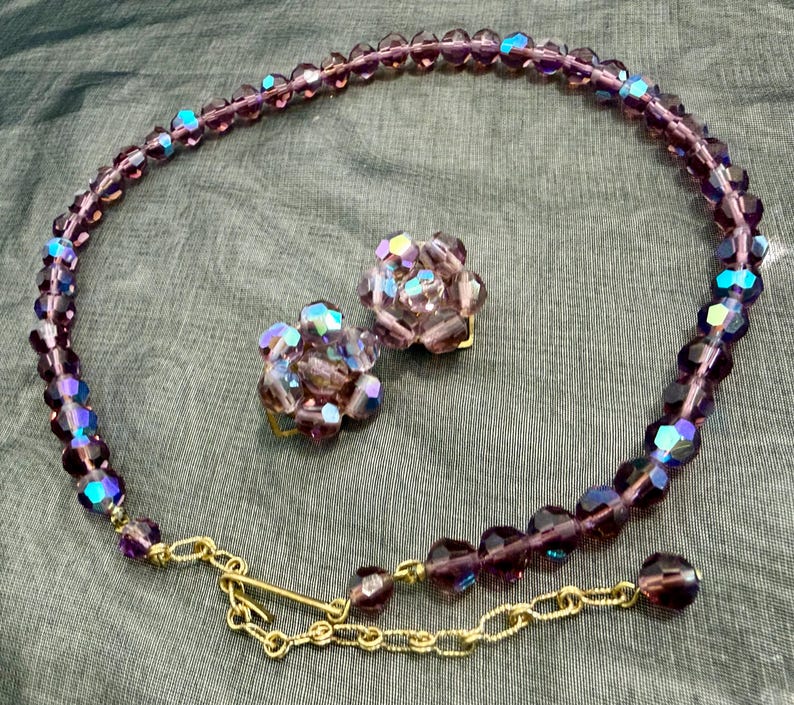 May include: A vintage jewelry set featuring a necklace and matching clip-on earrings. The necklace and earrings are made of faceted, iridescent purple beads. The necklace has a gold-tone chain and clasp.