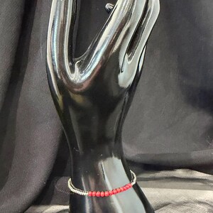 May include: A red beaded bracelet with a silver-colored band is displayed on a black hand-shaped jewelry holder. The bracelet features small, round red beads strung together. The jewelry holder is set against a dark background.