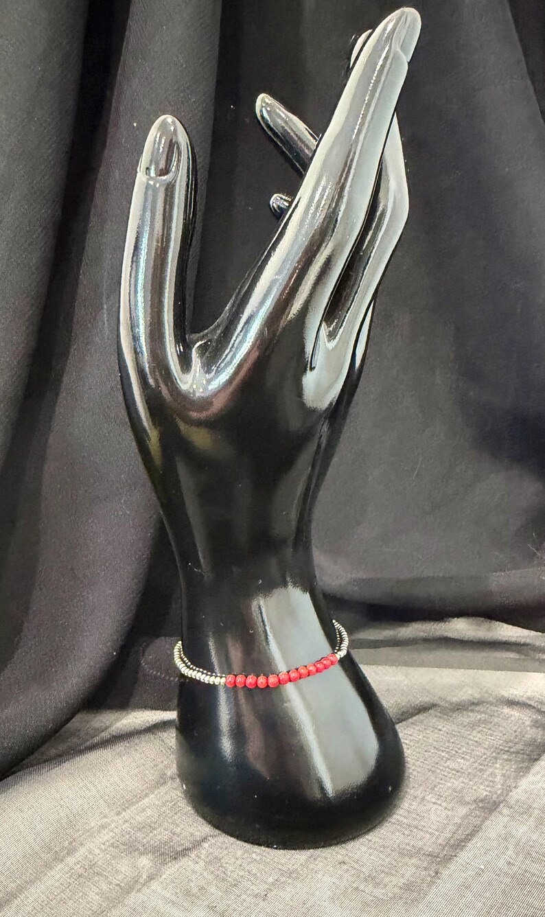 May include: A red beaded bracelet with a silver-colored band is displayed on a black hand-shaped jewelry holder. The bracelet features small, round red beads strung together. The jewelry holder is set against a dark background.