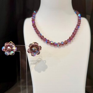 May include: A vintage jewelry set featuring a beaded necklace and matching clip-on earrings. The necklace and earrings are a deep purple color with iridescent accents. The earrings are flower-shaped.