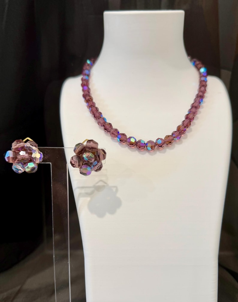May include: A vintage jewelry set featuring a beaded necklace and matching clip-on earrings. The necklace and earrings are a deep purple color with iridescent accents. The earrings are flower-shaped.