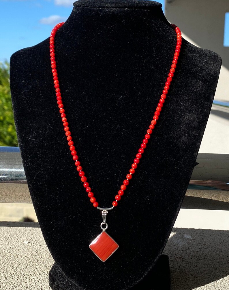 May include: A vibrant red beaded necklace with a diamond-shaped pendant. The pendant is a reddish-orange color, set in a silver-toned metal. The necklace is displayed on a black velvet bust against a sunny background.