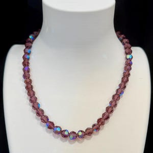 May include: A purple beaded necklace displayed on a white mannequin. The necklace features faceted beads with iridescent highlights. The beads are strung together to form a classic necklace design. The background is black.