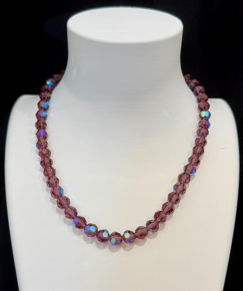 May include: A purple beaded necklace displayed on a white mannequin. The necklace features faceted beads with iridescent highlights. The beads are strung together to form a classic necklace design. The background is black.