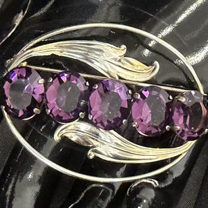 May include: An oval-shaped silver-tone brooch featuring five faceted, deep purple oval stones arranged in a horizontal row. The stones are framed by a polished silver-tone metal border with stylized leaf-like accents at the top and bottom.