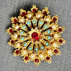 May include: A gold-toned brooch with a circular design. The brooch features a central red gemstone surrounded by turquoise, pearl, and red accents. The brooch has a detailed, ornate design, and is set against a textured gray background.