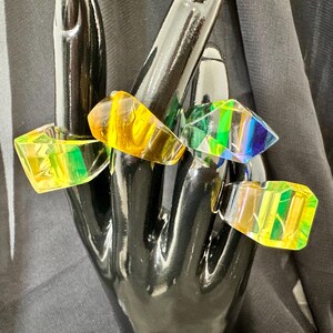 May include: Five colourful, geometric rings in shades of yellow, orange, green, and blue are displayed on a black hand-shaped display. The rings have a faceted design, creating a prism effect with light.