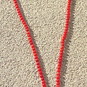 May include: A red beaded necklace with a silver clasp and a red square pendant. The necklace is made of small, round beads and is displayed against a textured, light brown surface.