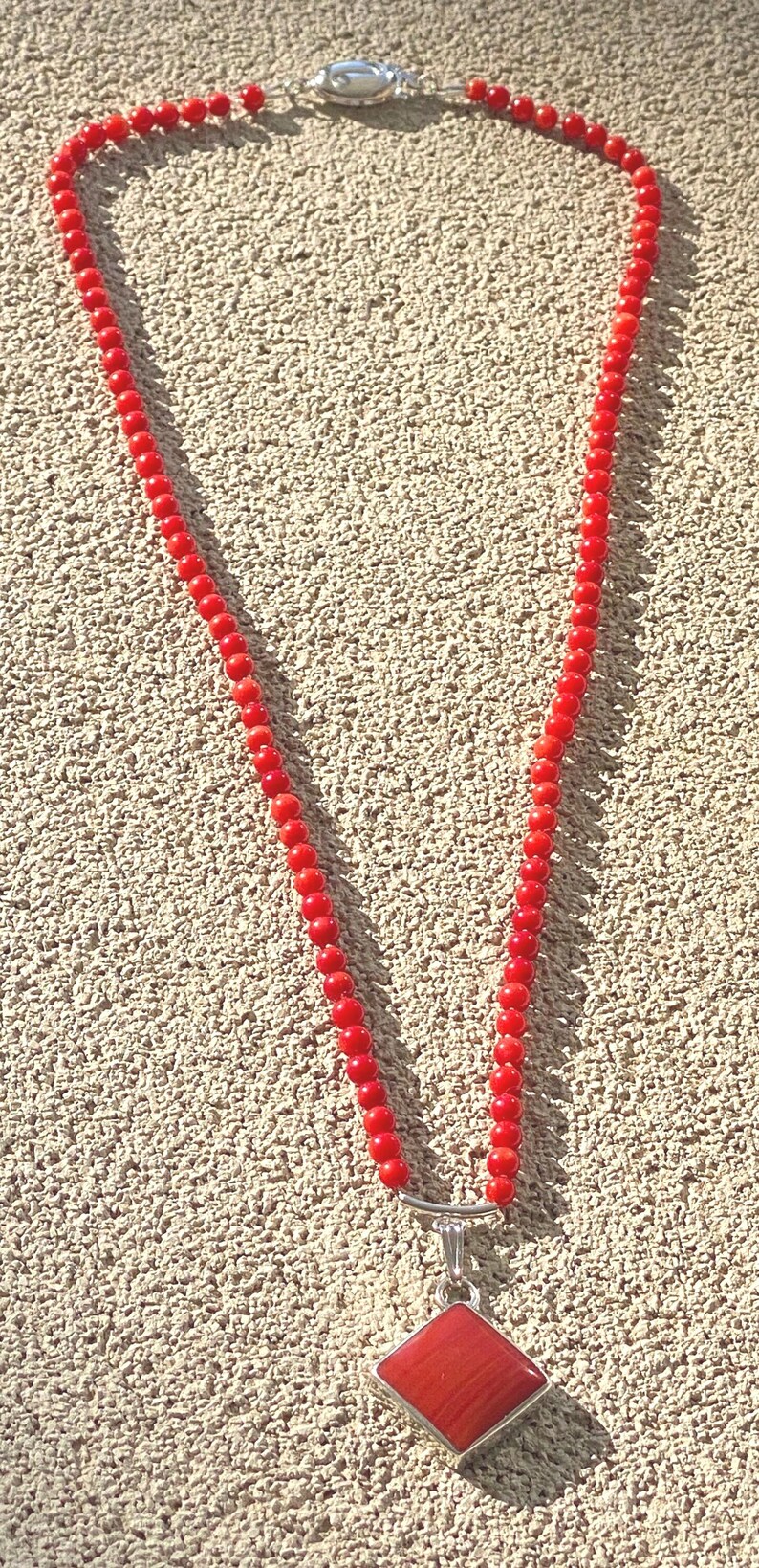 May include: A red beaded necklace with a silver clasp and a red square pendant. The necklace is made of small, round beads and is displayed against a textured, light brown surface.