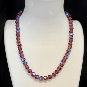 May include: A beaded necklace with faceted, purple beads. Some beads have an iridescent, blue-green sheen. The necklace is displayed on a white mannequin bust against a black background. A classic jewelry piece.