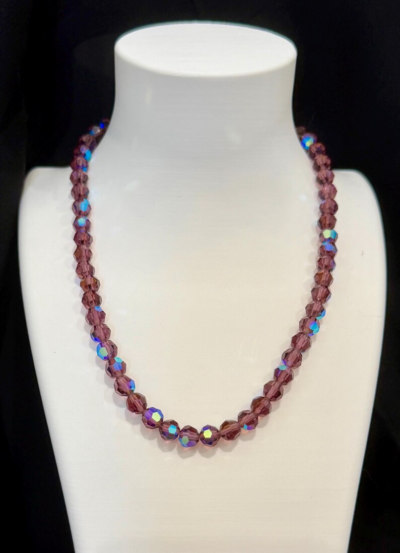 May include: A beaded necklace with faceted, purple beads. Some beads have an iridescent, blue-green sheen. The necklace is displayed on a white mannequin bust against a black background. A classic jewelry piece.