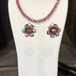 May include: A vintage jewelry set featuring a necklace and matching earrings. The necklace and earrings are made of faceted, iridescent purple beads. The earrings are flower-shaped. The jewelry is displayed on a white bust.