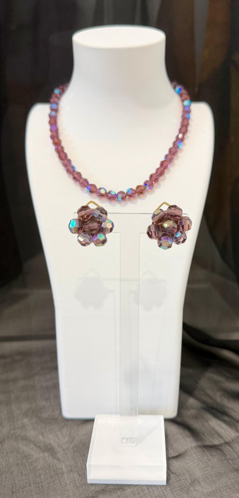 May include: A vintage jewelry set featuring a necklace and matching earrings. The necklace and earrings are made of faceted, iridescent purple beads. The earrings are flower-shaped. The jewelry is displayed on a white bust.