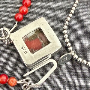 May include: A silver necklace with a square pendant featuring a red stone. The necklace has a beaded chain and a separate strand of red coral beads. The clasp and pendant are marked with "925".