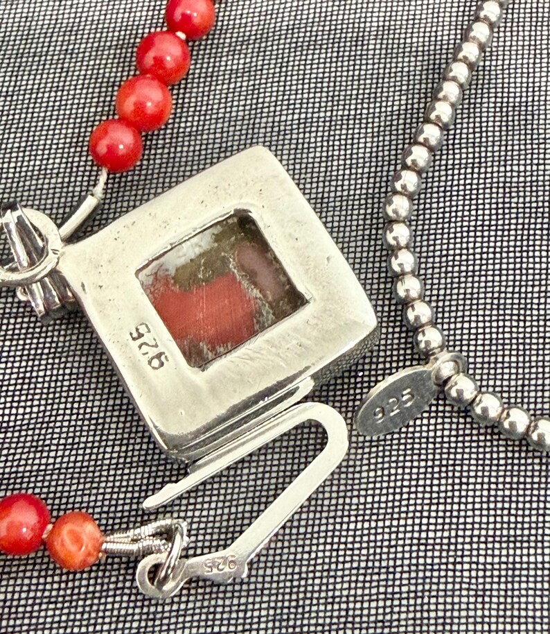 May include: A silver necklace with a square pendant featuring a red stone. The necklace has a beaded chain and a separate strand of red coral beads. The clasp and pendant are marked with "925".