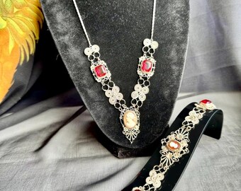 May include: A silver necklace and bracelet set featuring red and white cameo stones. The necklace has a V-shaped design with two red stones at the top and a cameo in the centre. The bracelet has a similar design with three red stones and a cameo.