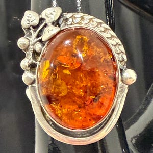 Könnte beinhalten: Ovale Bernsteinring mit Silberfassung. Der Bernsteinstein hat einen warmen, orangefarbenen Farbton mit inneren Mustern. Das Silberband weist ein dekoratives Blatt- und Weinreben-Design auf und verleiht dem Schmuck einen Hauch von von der Natur inspirierten Eleganz.
