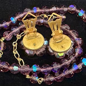 May include: A vintage jewelry set featuring a necklace and clip-on earrings. The necklace and earrings are made of faceted, purple beads with iridescent highlights. The earrings have gold-toned metal clips. The set is displayed on a black surface.