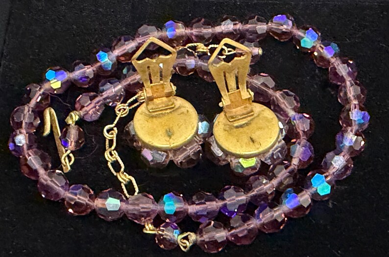 May include: A vintage jewelry set featuring a necklace and clip-on earrings. The necklace and earrings are made of faceted, purple beads with iridescent highlights. The earrings have gold-toned metal clips. The set is displayed on a black surface.