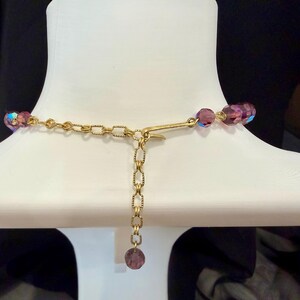May include: A close-up of a necklace on a white mannequin. The necklace features iridescent purple beads and a gold-colored chain. The clasp and extender chain are visible, with a single purple bead at the end of the extender.