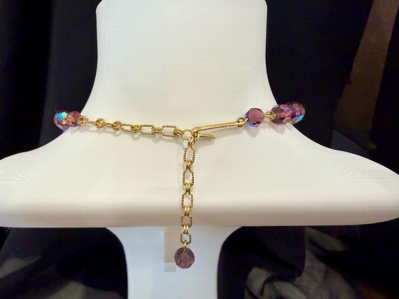 May include: A close-up of a necklace on a white mannequin. The necklace features iridescent purple beads and a gold-colored chain. The clasp and extender chain are visible, with a single purple bead at the end of the extender.