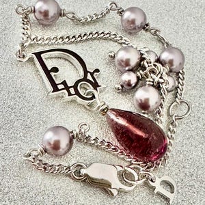 May include: A silver-toned bracelet featuring a 'Dior' charm, lavender-colored faux pearls, and a teardrop-shaped, deep red glass bead. The bracelet has a chain link design and a clasp closure.