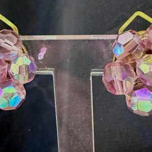 May include: A pair of clip-on earrings featuring clusters of faceted, iridescent pink beads. Each cluster is attached to a small, gold-colored metal backing. The earrings have a vintage aesthetic and are displayed against a dark background.