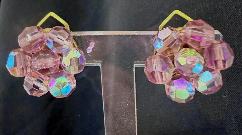 May include: A pair of clip-on earrings featuring clusters of faceted, iridescent pink beads. Each cluster is attached to a small, gold-colored metal backing. The earrings have a vintage aesthetic and are displayed against a dark background.
