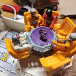 Op de afbeelding: Vier astronaut-figuurtjes zitten rond een paarse en grijze tafel met een paars middenstuk. De astronauten dragen witte, gele en rode ruimtepakken. De tafel is omringd door vier gele stoelen.