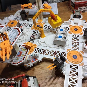 Könnte beinhalten: Ein weiß-oranges Raumstationsspielzeug mit Kränen, Modulen und Figuren. Das Spielzeug hat orangefarbene und schwarze Akzente, wobei das Wort "WEIGHT" sichtbar ist. Das Spielzeug befindet sich auf einer Holzoberfläche.