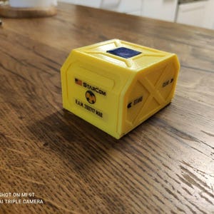 Op de afbeelding: Een gele, rechthoekige doos met een blauw vierkantje bovenop. Op de doos staat de tekst "STARCOM" en "R.A.M. 260717 MAB", samen met een gevaarsymbool. De doos staat op een houten oppervlak.