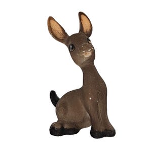 Puede incluir: Pequeña figura de burro marrón con grandes orejas color canela y cola negra. El burro está sentado, con la cabeza ligeramente inclinada y una expresión sonriente. La figura parece estar hecha de cerámica o material similar.
