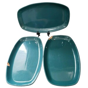 May include: Three teal ceramic serving platters with smooth, glossy finishes and rounded rectangular shapes. One platter is positioned horizontally on a black stand, while two others are placed vertically below, showing small chips on the edges.